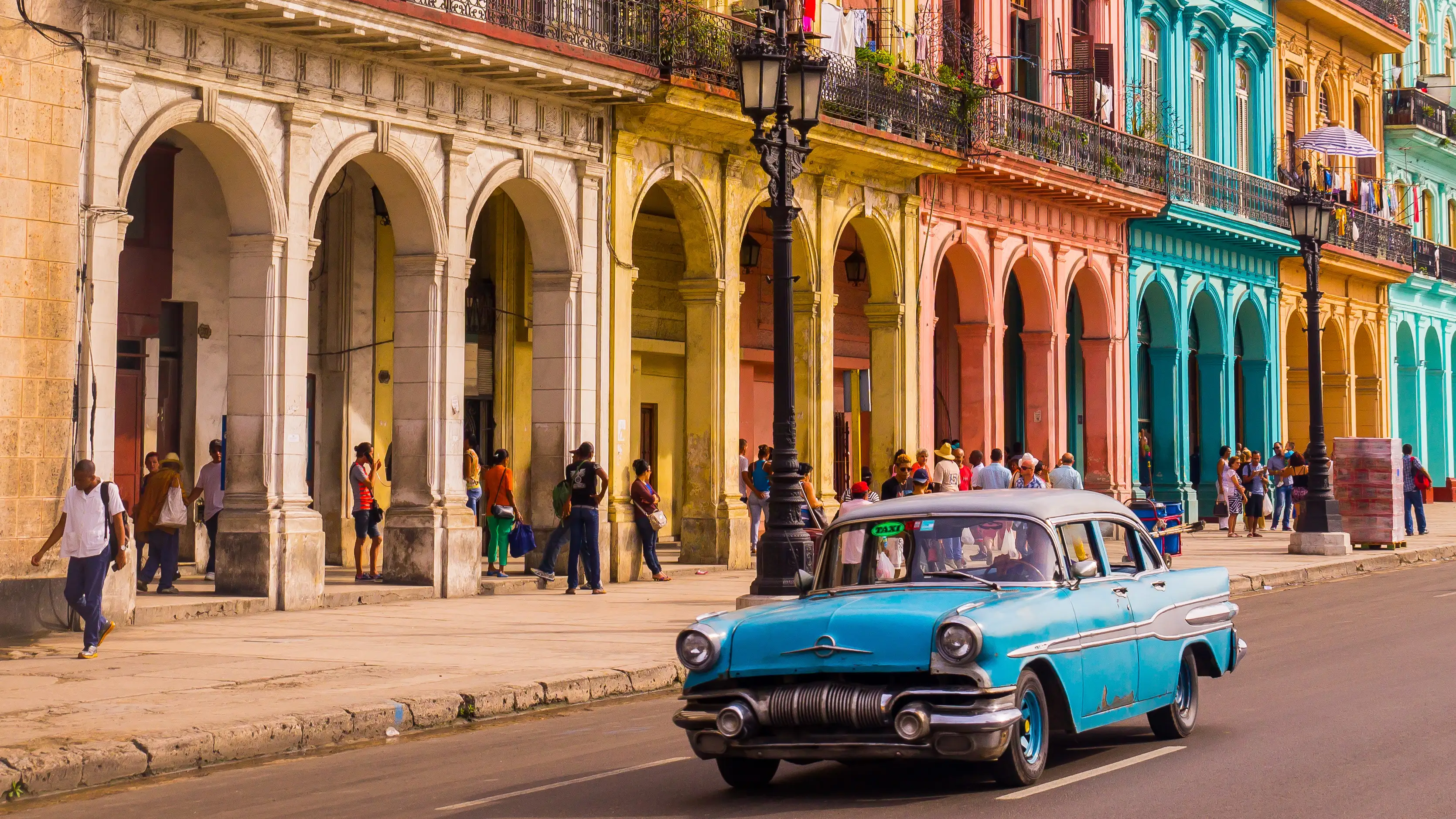 A blue oldtimer taxi is driving through Habana Vieja in front of a colorful façade.