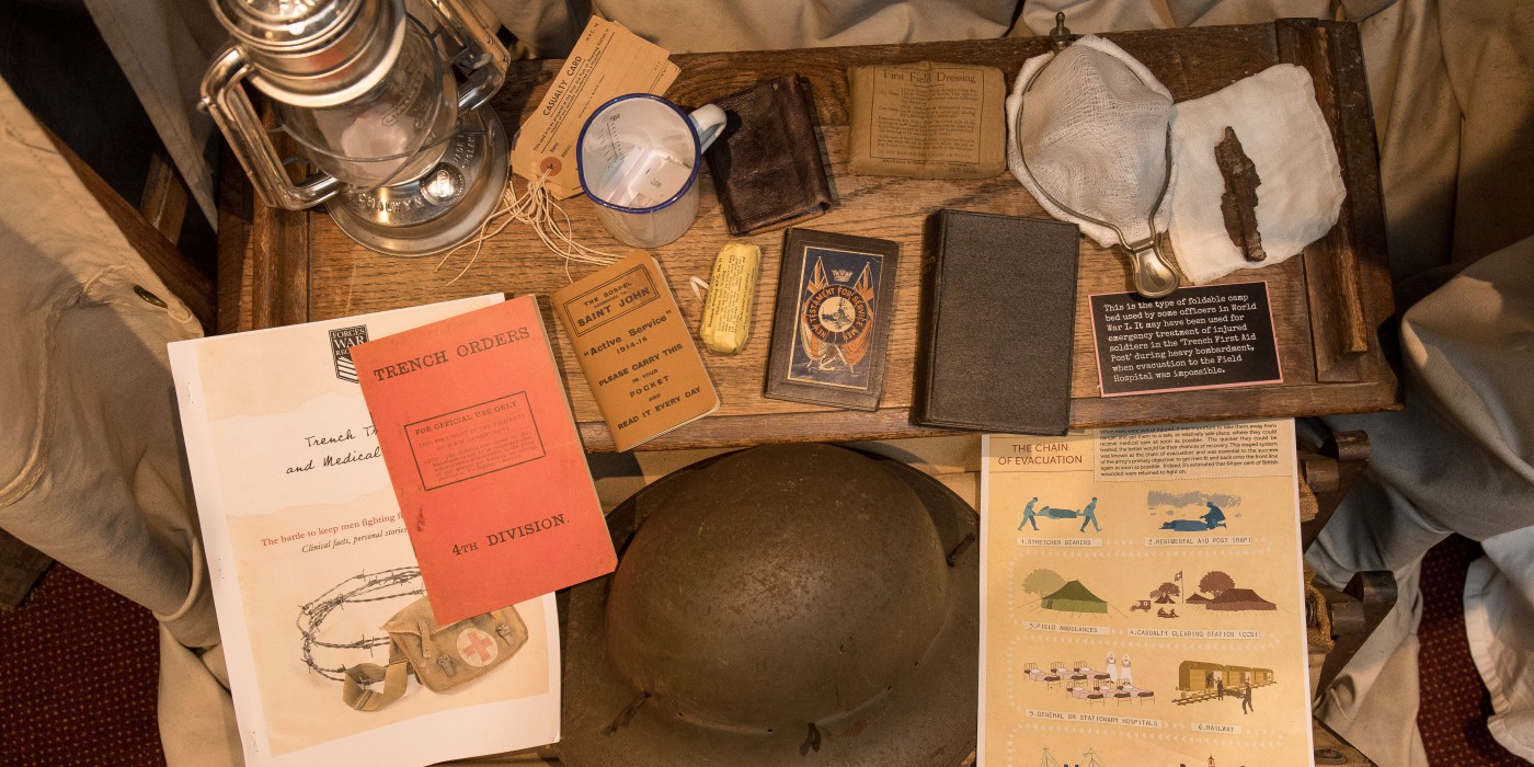 Various items found with a soldier, including a lantern, trench orders booklet, mirror, soap, mug, casualty cards, a variety of books and guides, field dressing, all of the items are placed on top of a wooden foldable camp bed used by some officers in WW1.