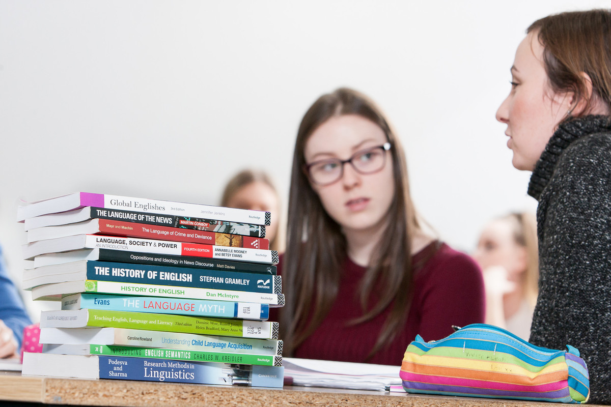 Two female students itting at a table talking with a stack of English language books.