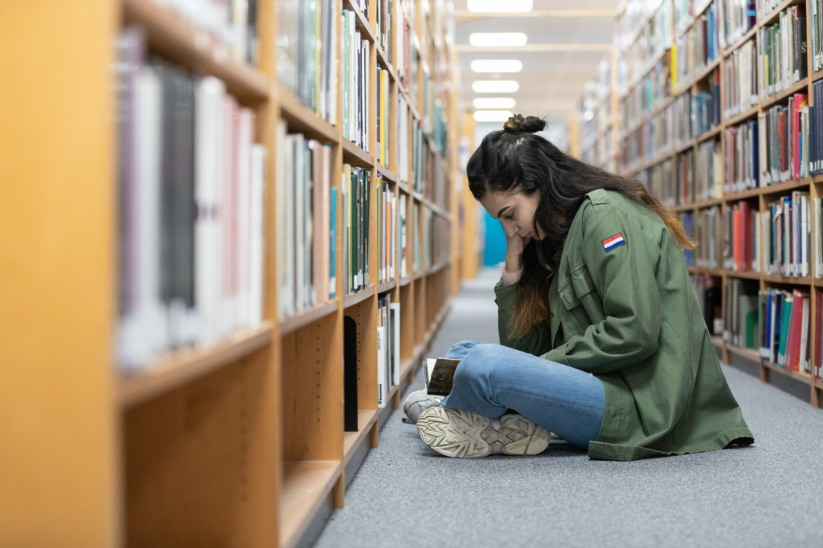 A student sat on the floor in a library reading a book