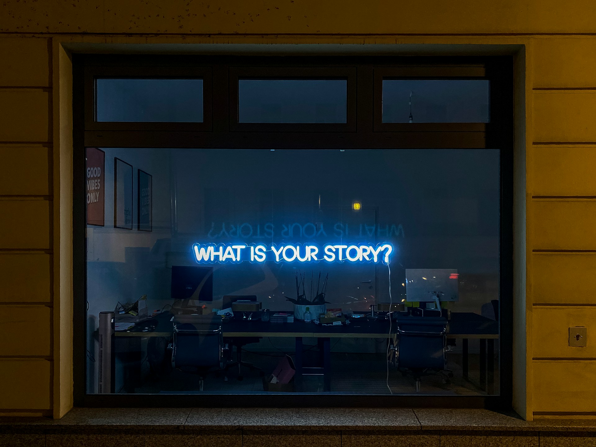 A neon sign saying what is your story, hanging in a window at night