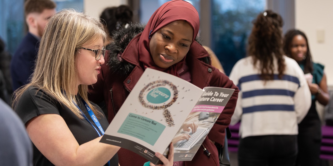 A member of staff and student at an event discussing and looking at a Guide to Your Future Career. With other people blurred in the background.