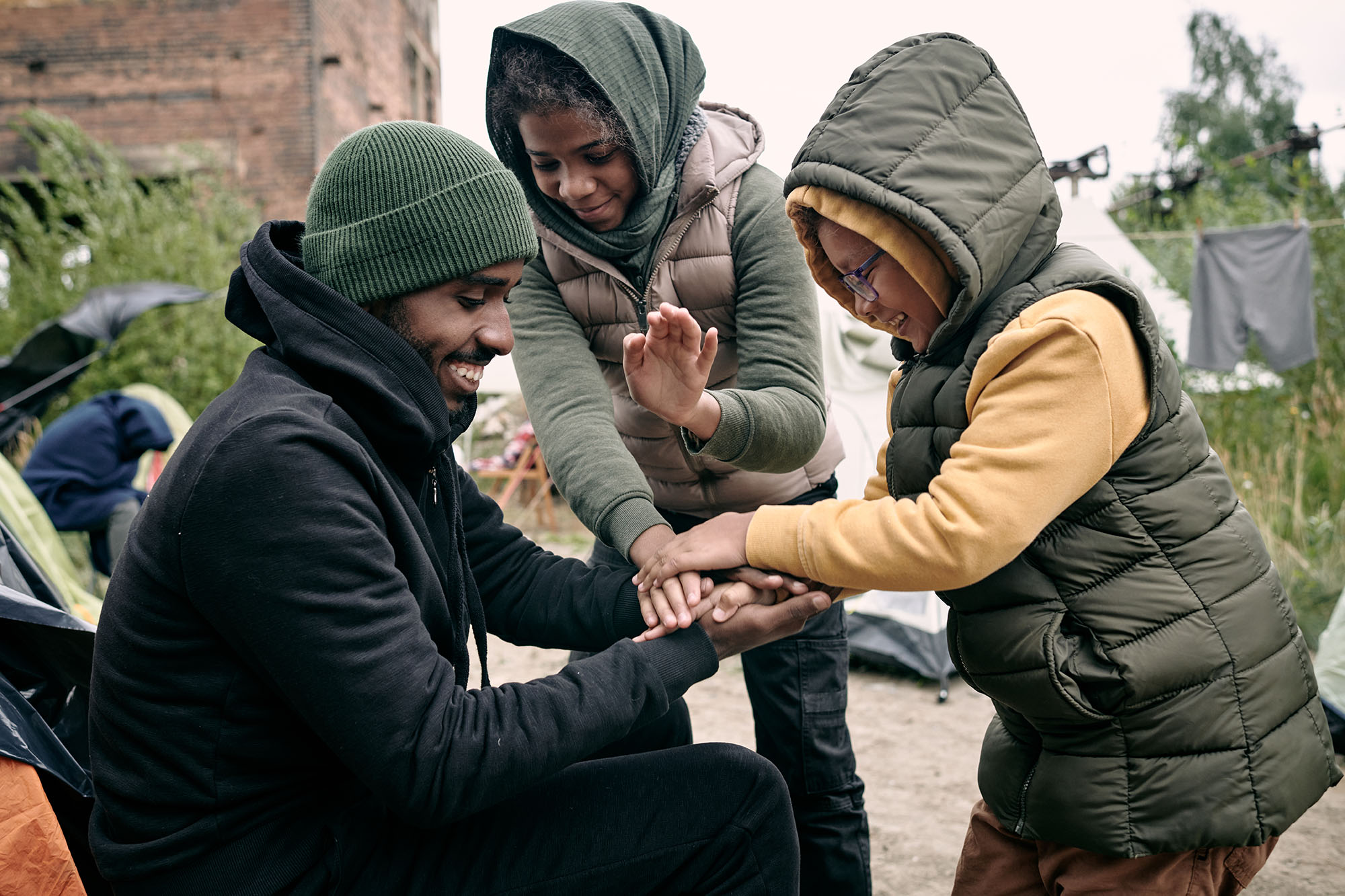 Positive young Black man in hat playing with kids against abandoned building and hanging clothes on rope in refugee camp