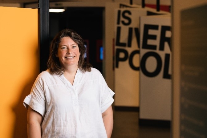 Laura Pye (Director of National Museums Liverpool) standing, facing the camera, smiling. In the background a sign that states Is Liverpool.