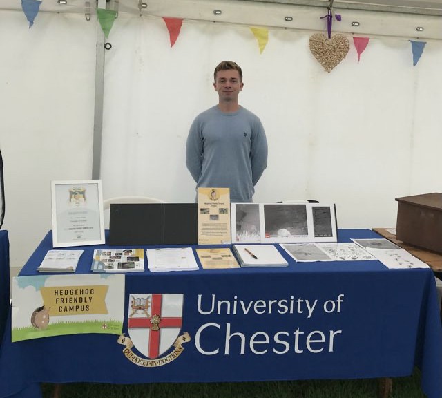 Lyndon Howson (student) standing behind a pop-up table. Looking at the camera and smiling. University of Chester Logo on table-cloth draped over table.