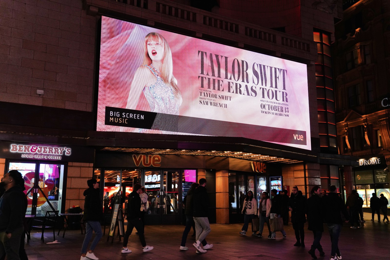 Taylor Swift Billboard's advertised outside a VUE building. People walking in both directions past the billboard in the evening.