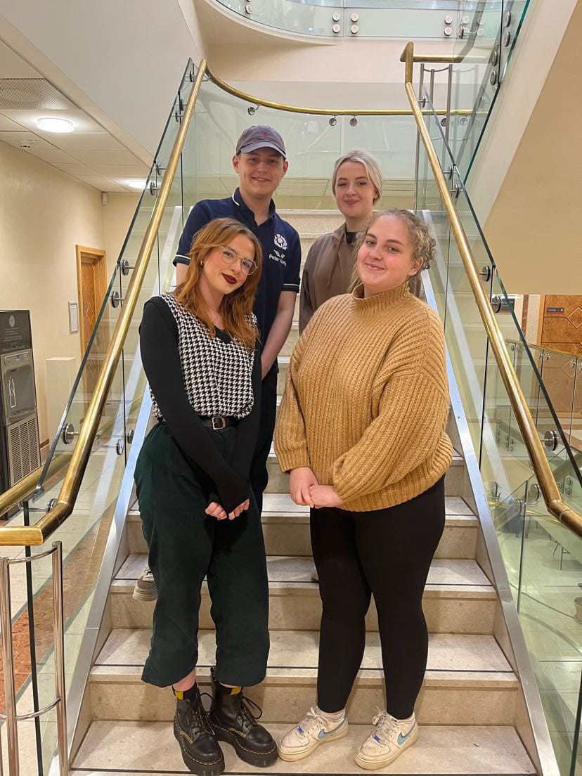 Four students standing on stairs, posing, facing the camera and smiling whilst standing on stairs in Queen's Park, Chester campus.