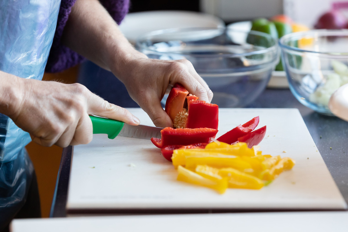 A pair of gloved hands, using a knife to cut red and yellow peppers on a chopping board, with glass bowls on a kitchen worktop.