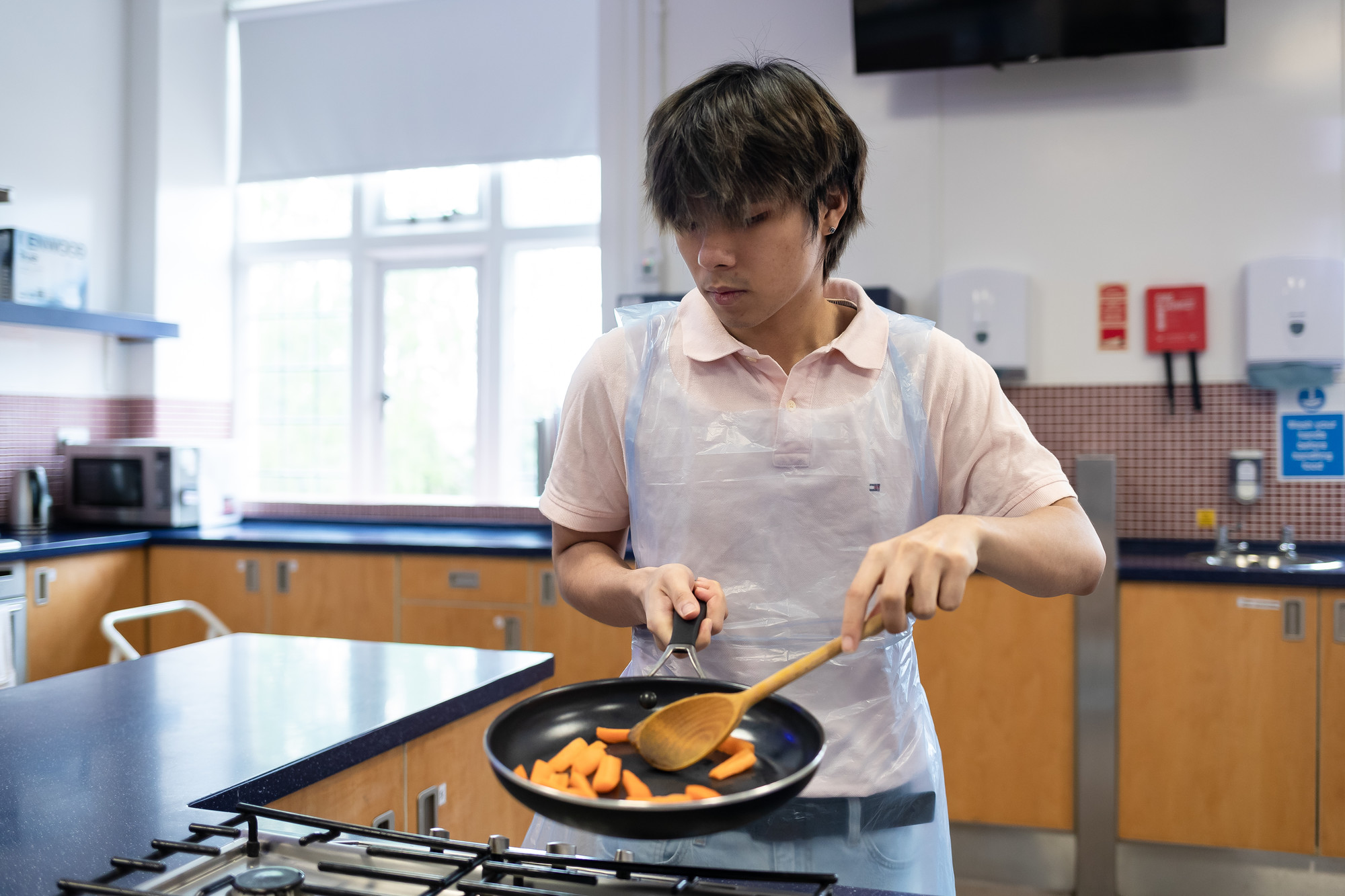 Student cooking a meal, holding a wooden spoon with one hand and a frying pan with the other