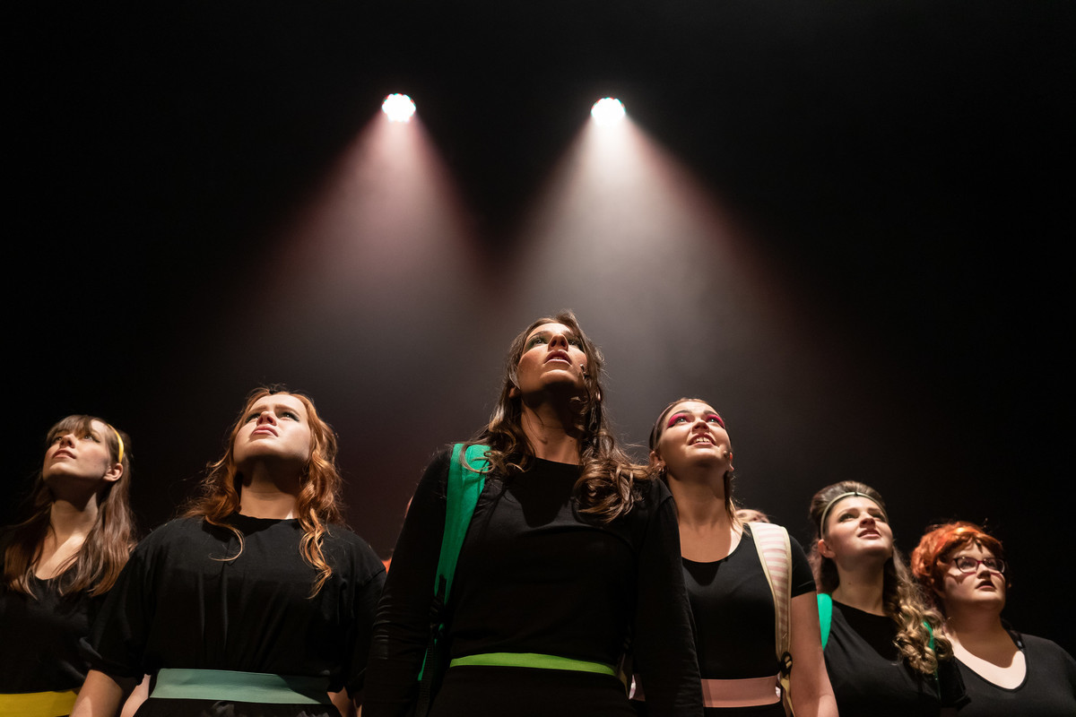 A group of students learning to perform musical theatre under two lit stage spotlights where the background is dark.
