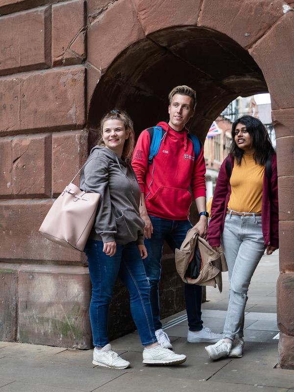 Two female and one male student walking in the street past city centre brick building. 