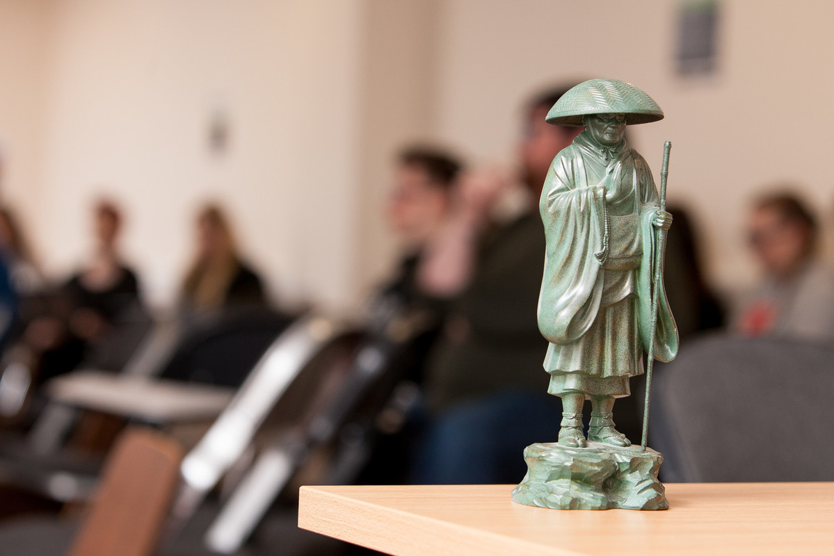 Students sitting in row seats in a lecture room with a green statute on table top.